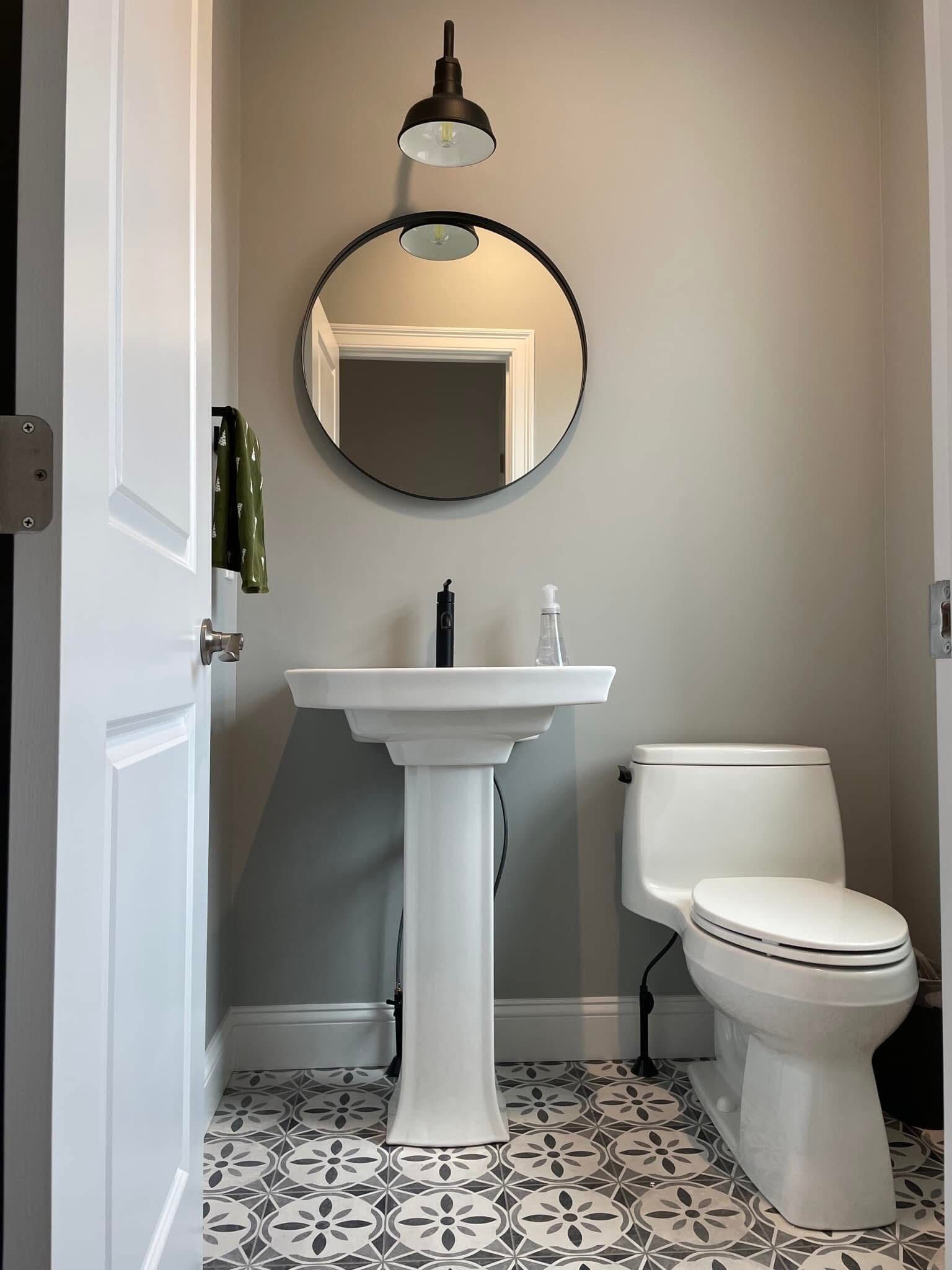 Small, white-walled bathroom with patterned floor, pedestal sink, round mirror, and toilet. A black light fixture hangs above the mirror.