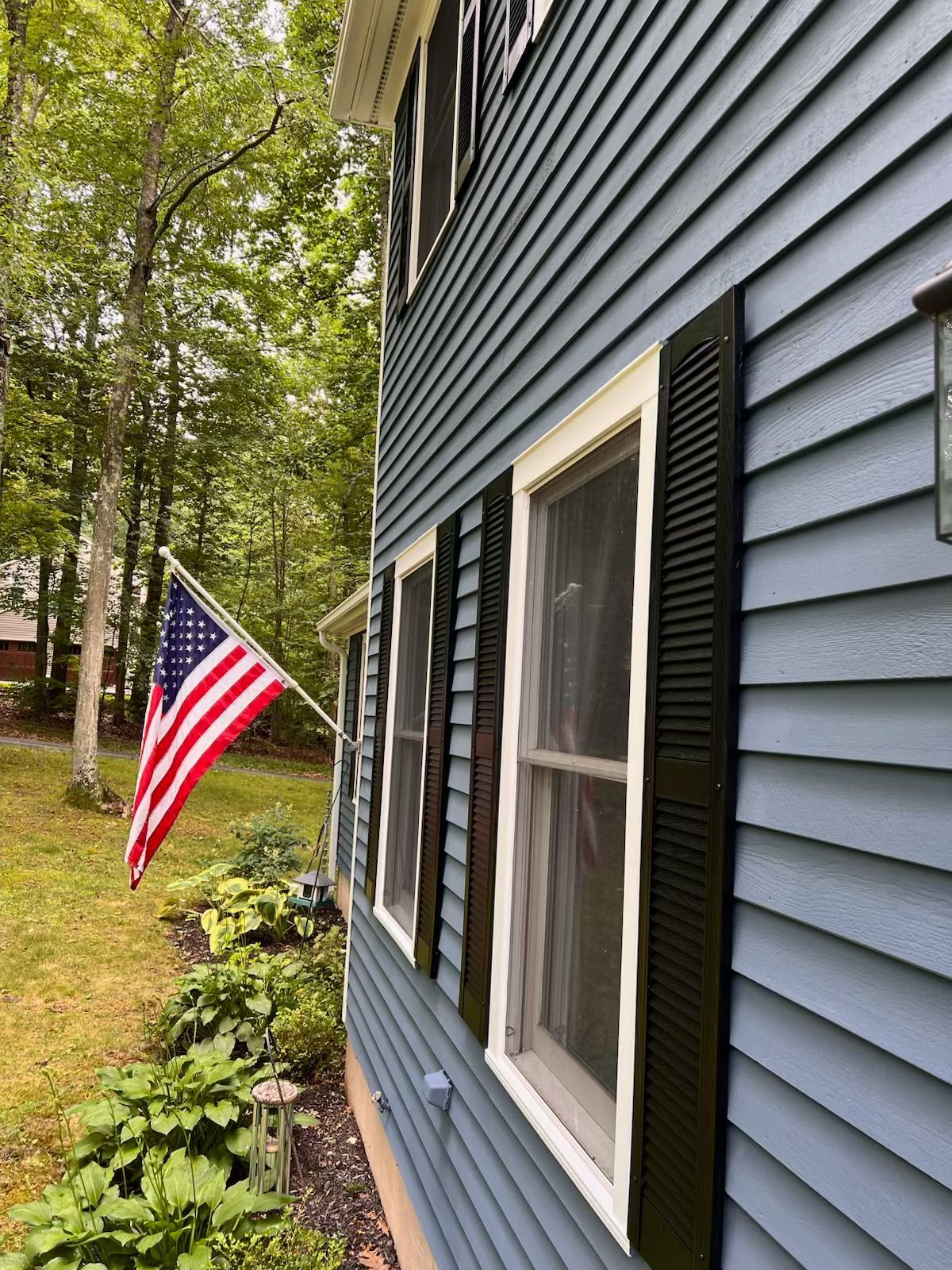 Side view of a two-story house with blue siding, white trim, and black shutters. An American flag waves in the front yard.