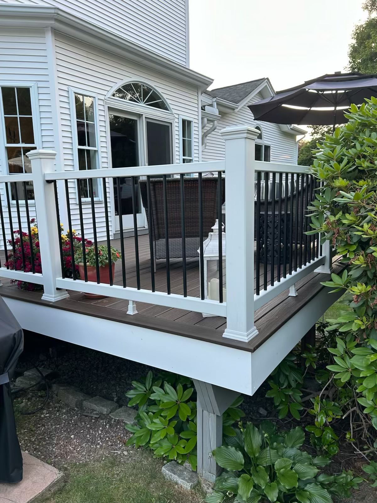 A white deck with black railings adjoins a white house. Potted flowers and outdoor furniture are visible.