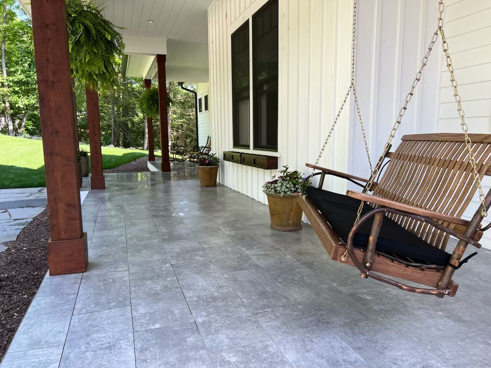 A porch with a gray tile floor, white siding, and a wooden swing. Brown columns support the roof, and there are potted plants.