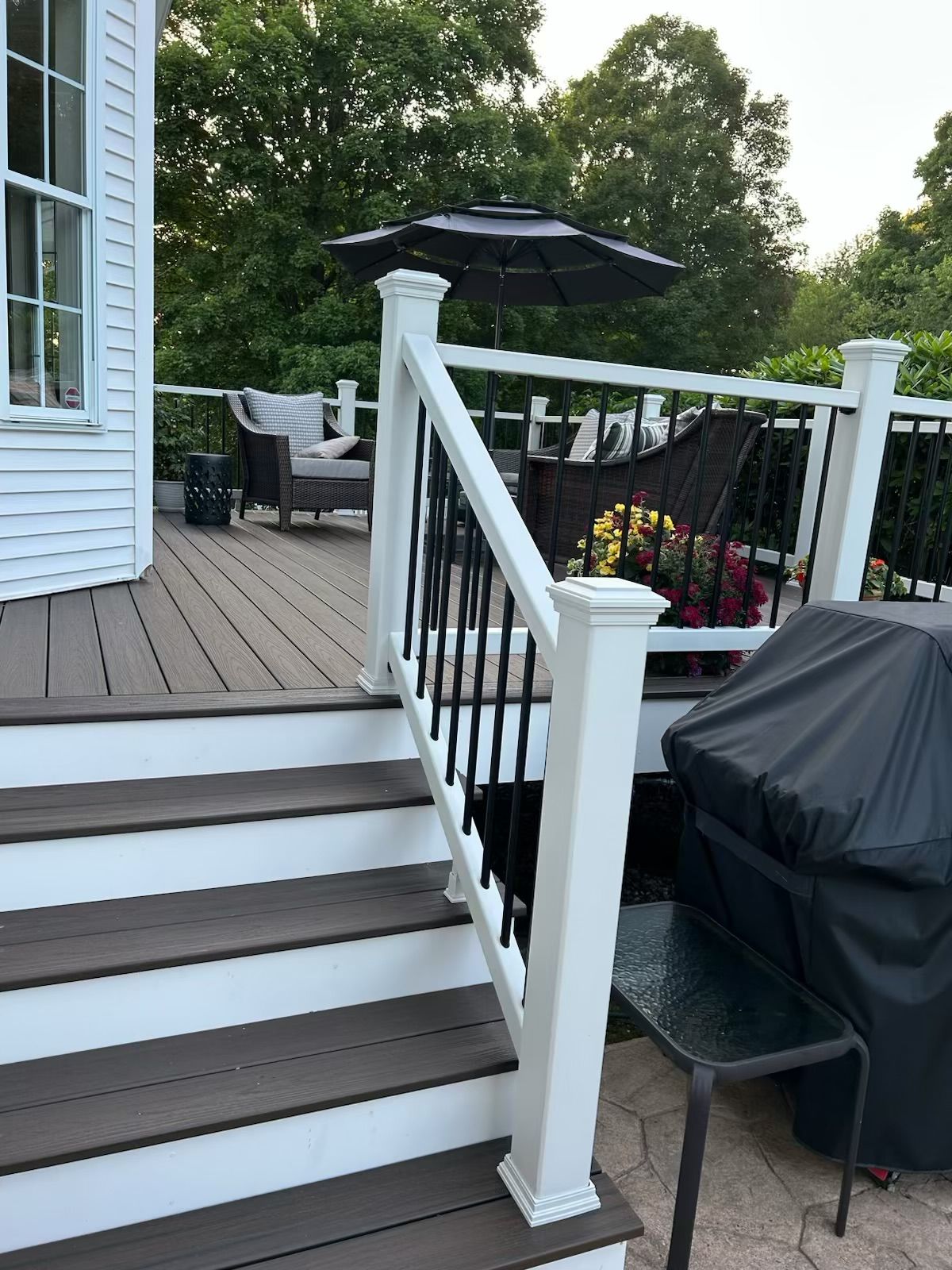 A deck with dark brown steps and railing, white posts, and black spindles. Outdoor seating and a grill are visible.