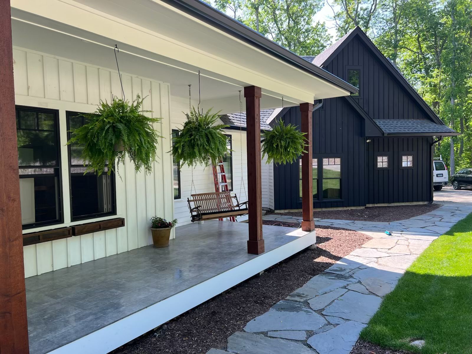 White farmhouse with a porch, black trim and hanging plants. A black building with windows sits next to the farmhouse.
