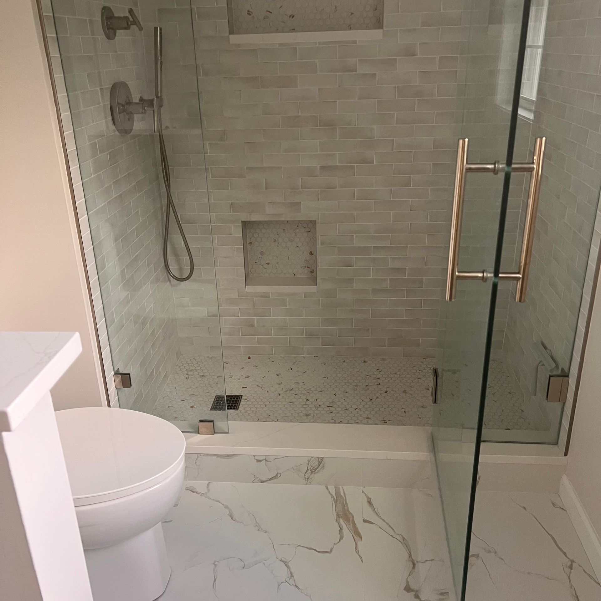 Bathroom with a glass-enclosed shower, white toilet, and marble flooring. The shower has a tiled wall with a built-in niche.