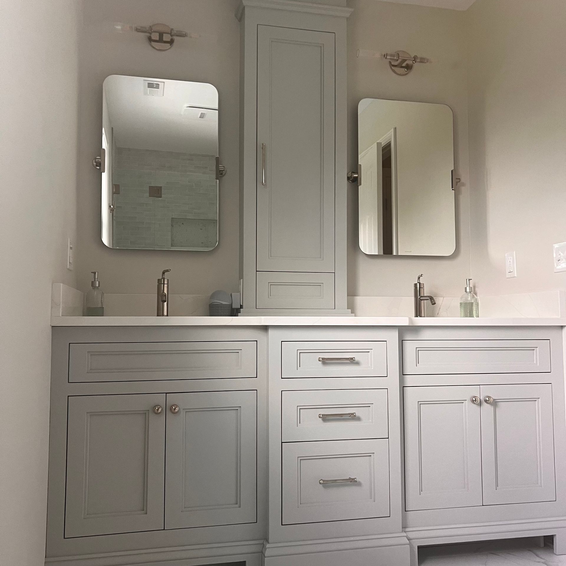 A bathroom with light gray cabinets, two mirrors, and a tall cabinet in the center. Sinks, faucets, and wall sconces are visible.