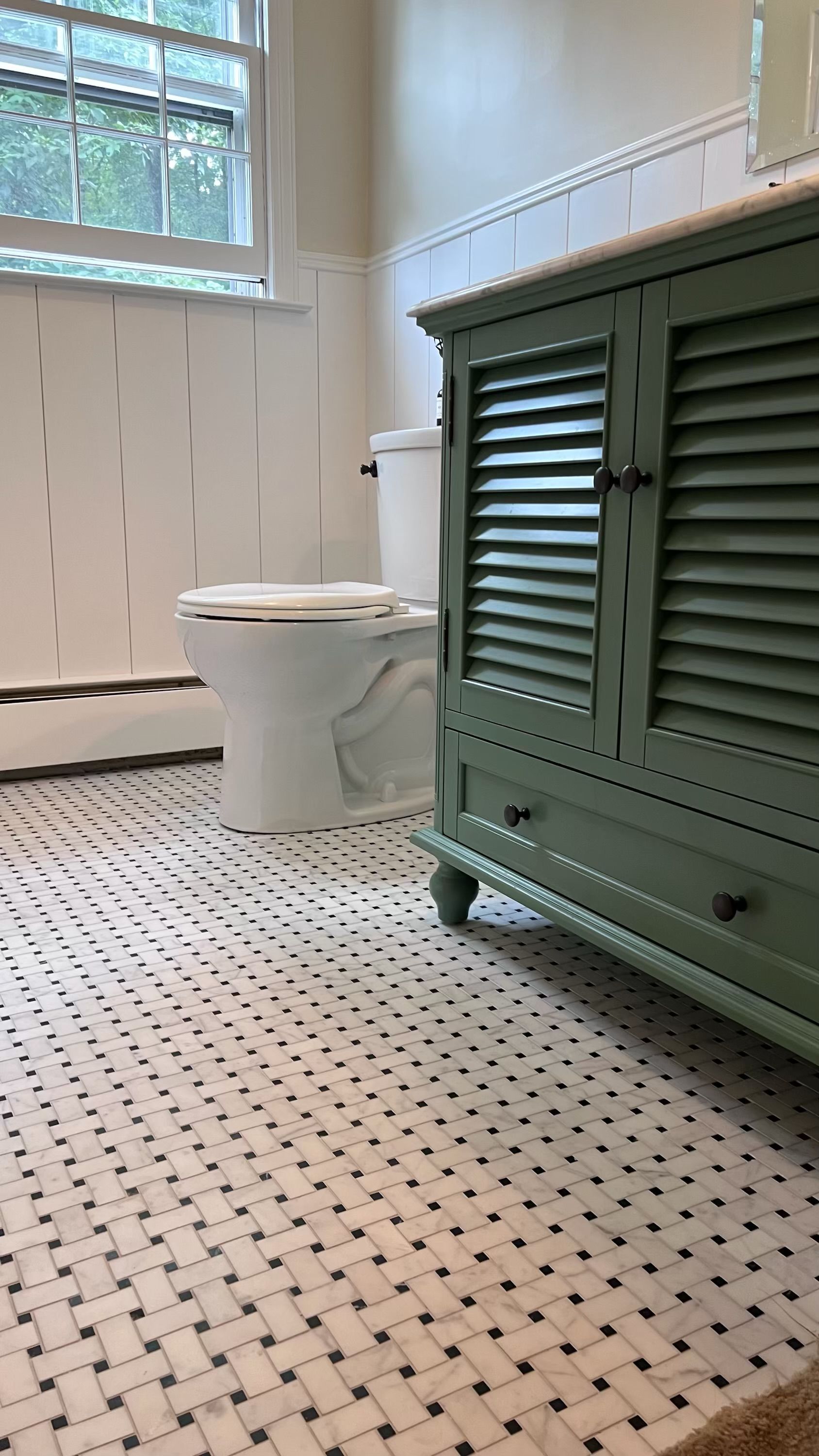 Bathroom with white and black tile floor, white toilet, and a green shutter-style cabinet. White paneling on the wall.