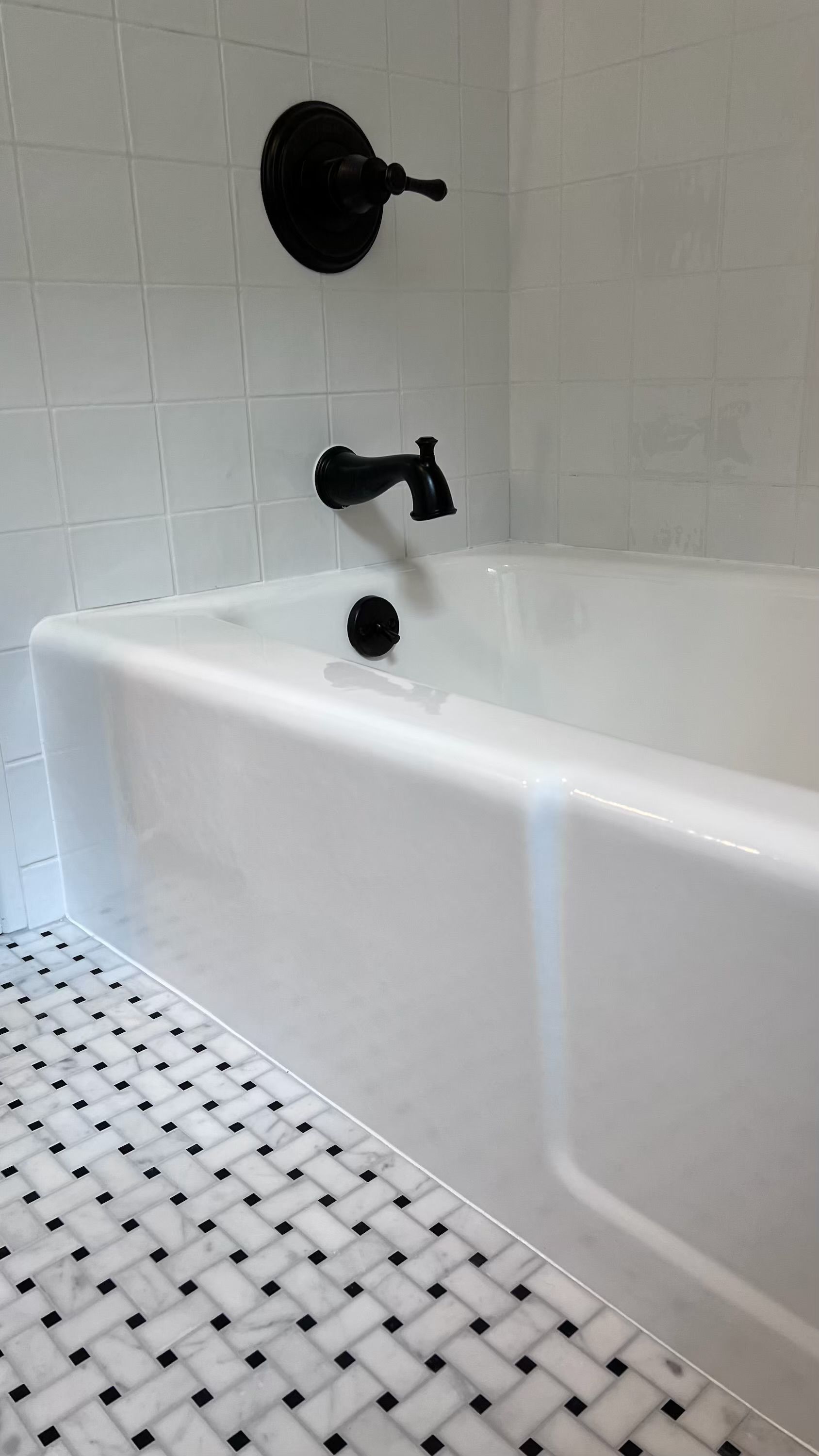 White bathtub with black fixtures against white tiled wall, set on marble floor with black and white tiles.