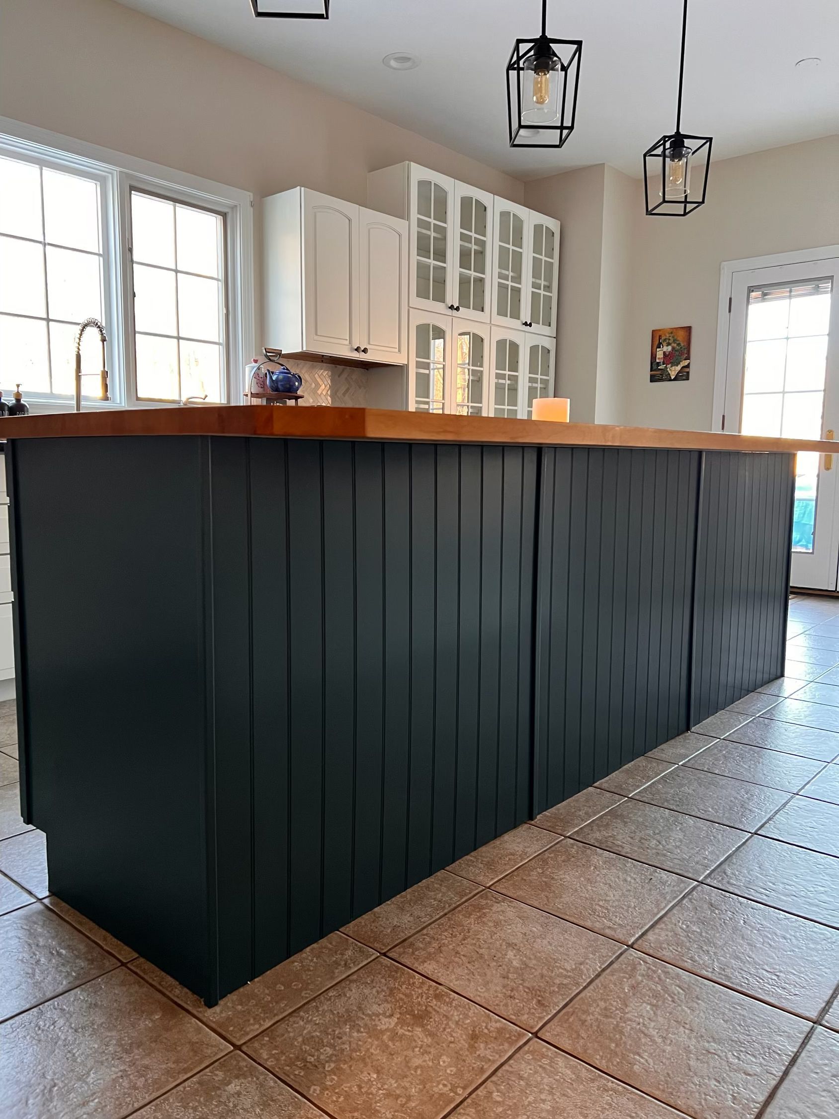 Dark green kitchen island with wood countertop, in a bright kitchen with white cabinets and two hanging pendant lights.