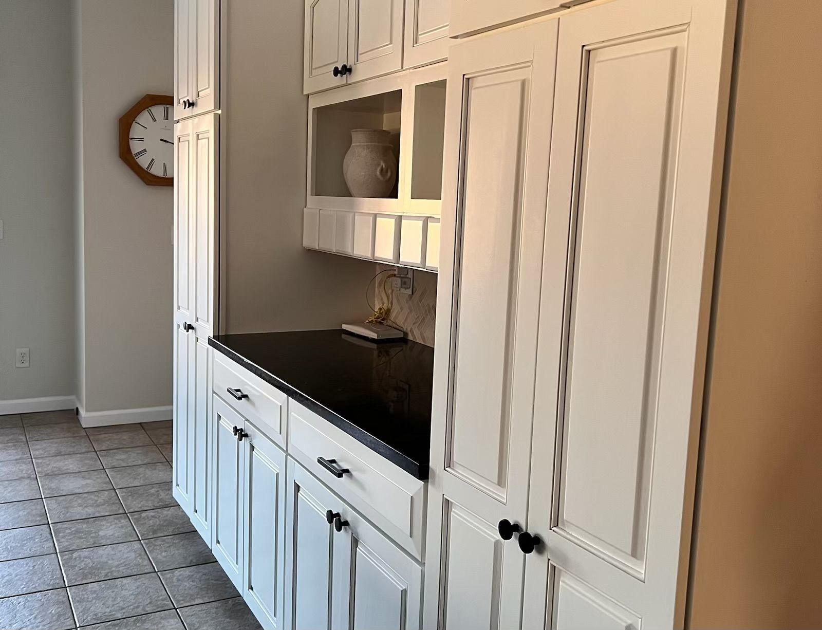 White kitchen cabinets with black countertops, clock on wall, and tile flooring.