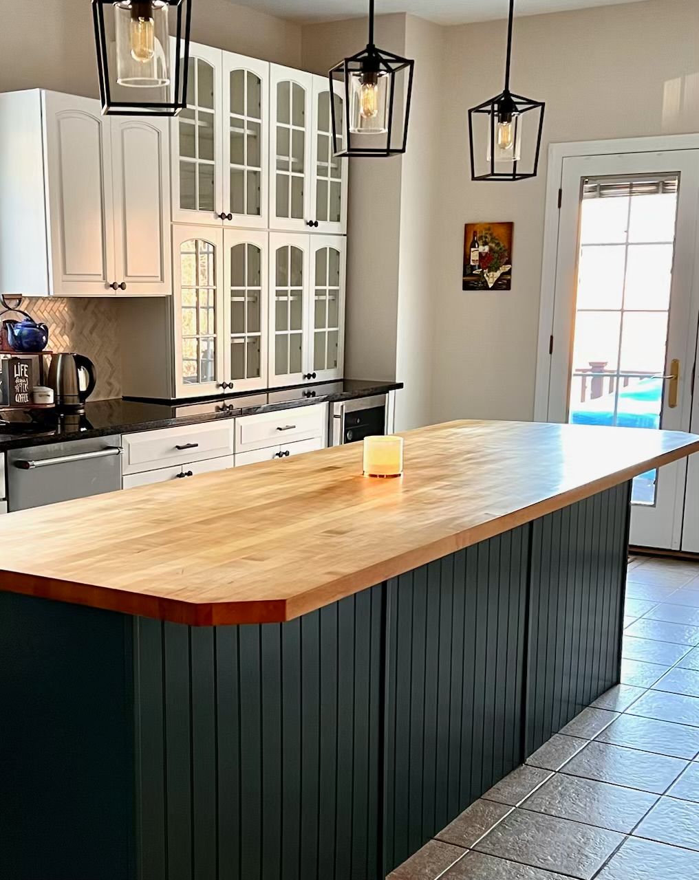 A kitchen with a large wooden island with a dark green base, white cabinets, and dark pendant lights.