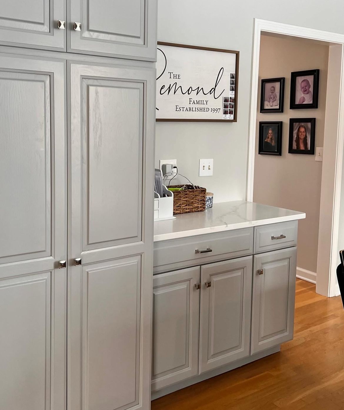 Gray kitchen cabinets with a white countertop, a sign, and framed photos.
