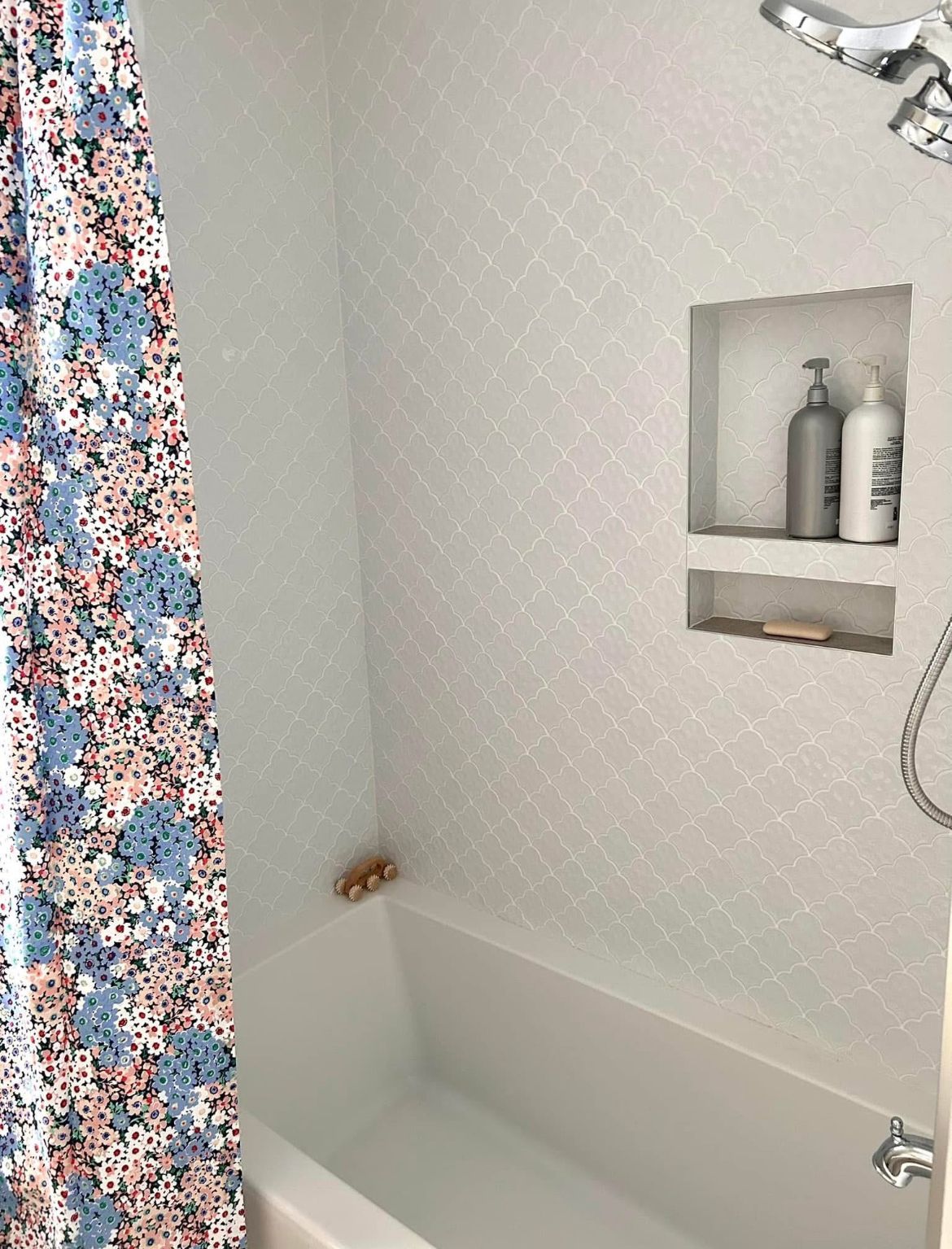 A bright white bathroom with patterned tile, a recessed shelf holding bottles, and a floral shower curtain.