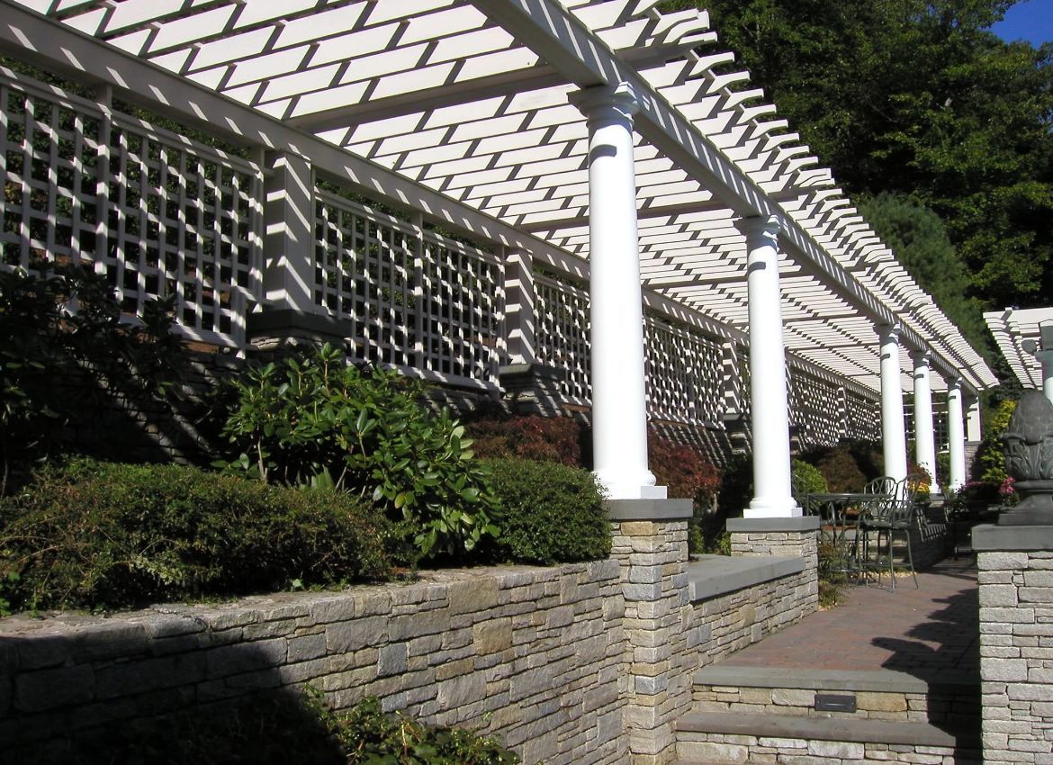White pergola with lattice and columns over a stone wall with shrubbery.