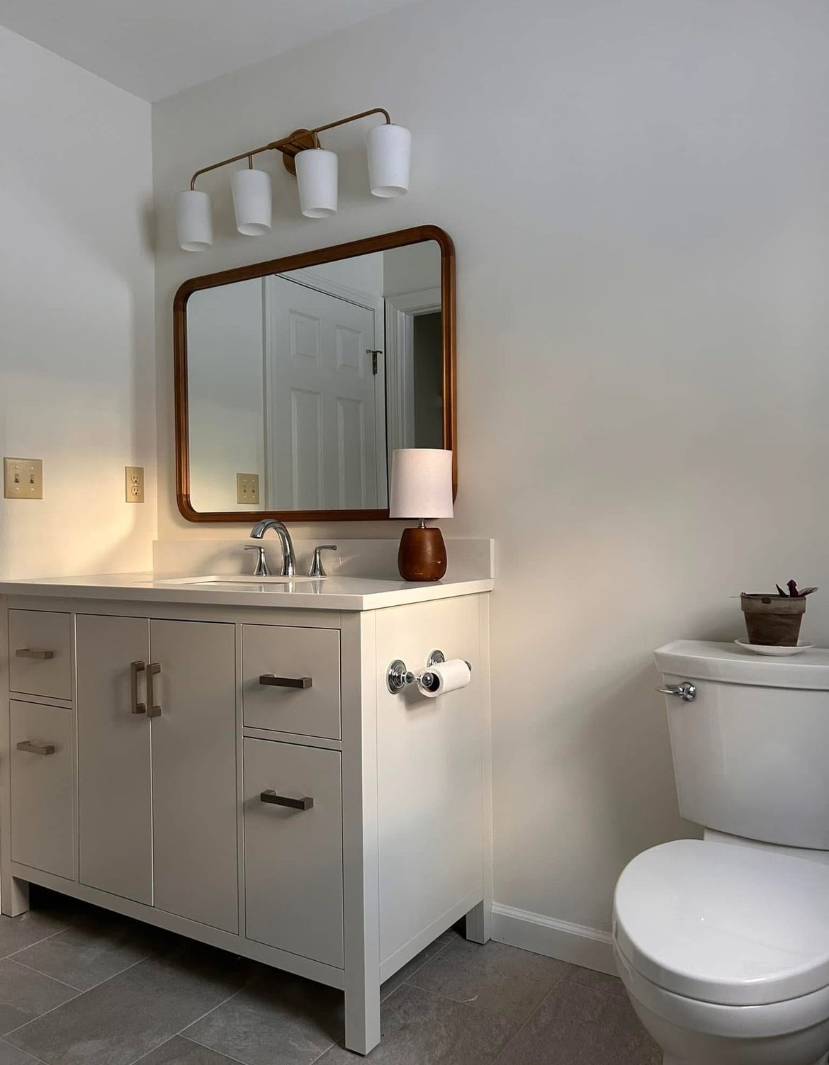 Bathroom with white vanity, wooden-framed mirror, and toilet. A small lamp sits on the counter, and a light fixture above the mirror.