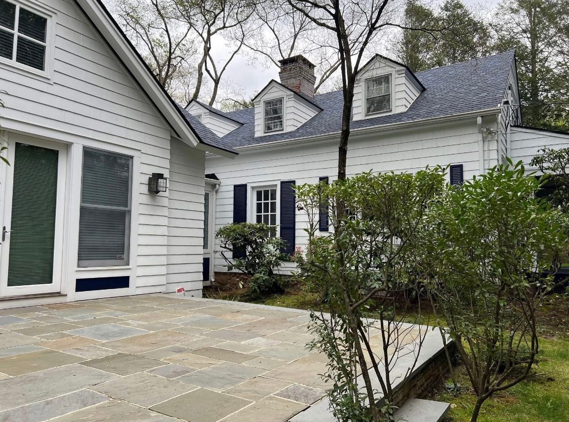 White house with blue shutters and a stone patio. Green bushes and trees are in the foreground.