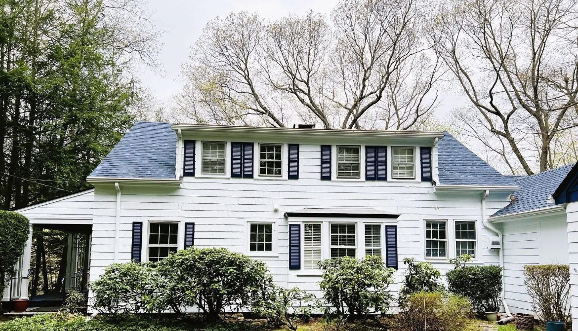 White house with blue roof and shutters, surrounded by trees and bushes.