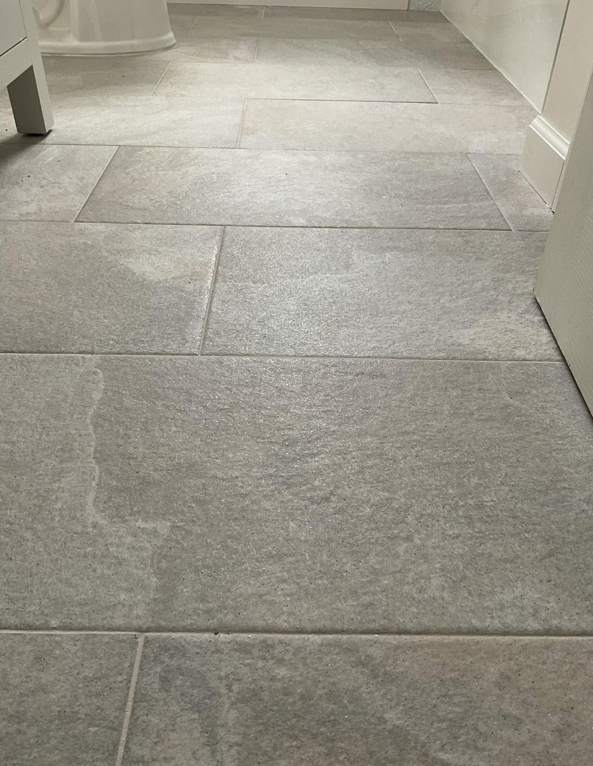 Gray rectangular stone floor tiles in a bathroom, seen from a low angle.  White trim and the base of a cabinet are visible.