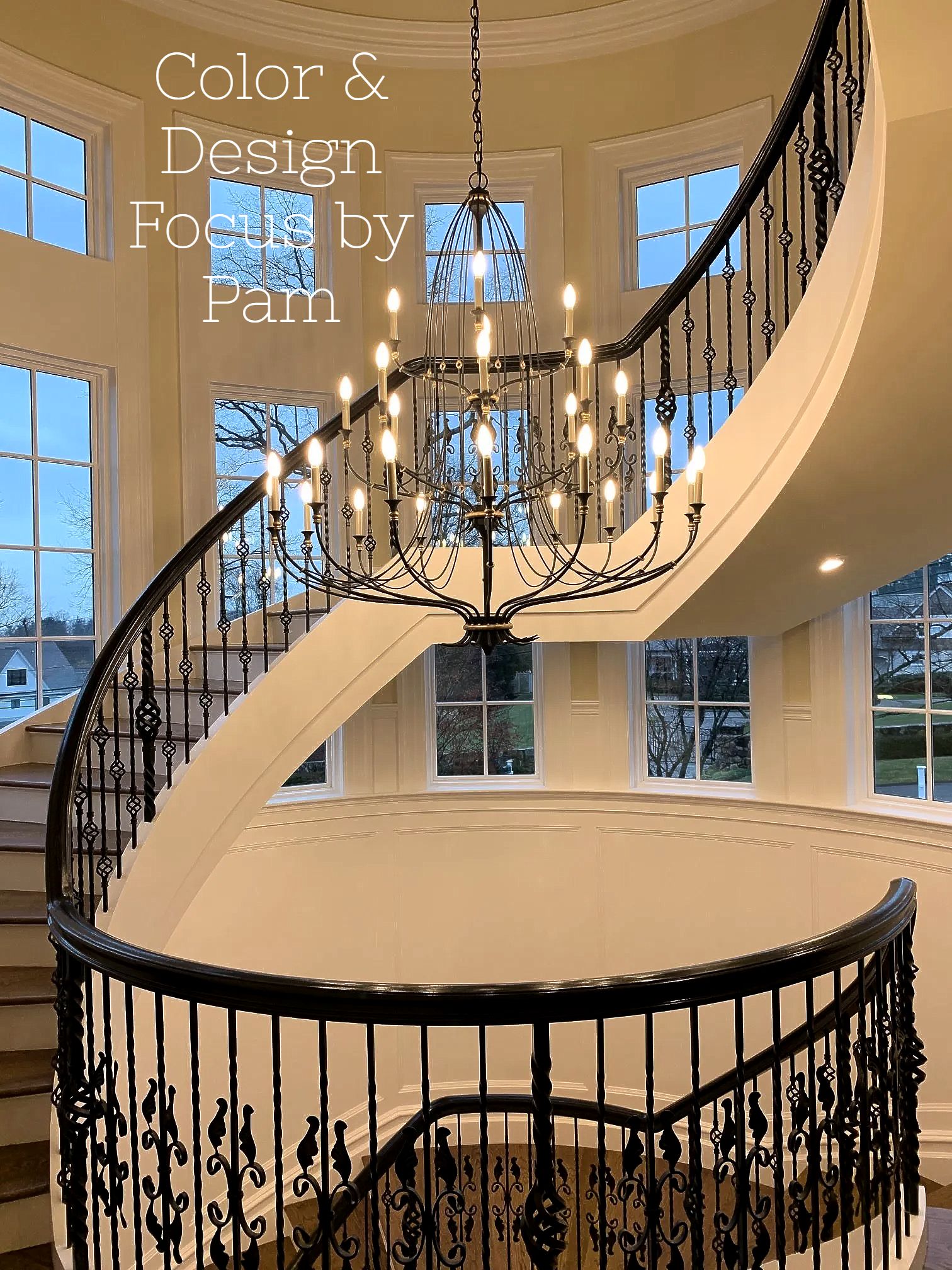 Elegant spiral staircase with black wrought iron railing and a large chandelier. Windows line the walls with outdoor views.