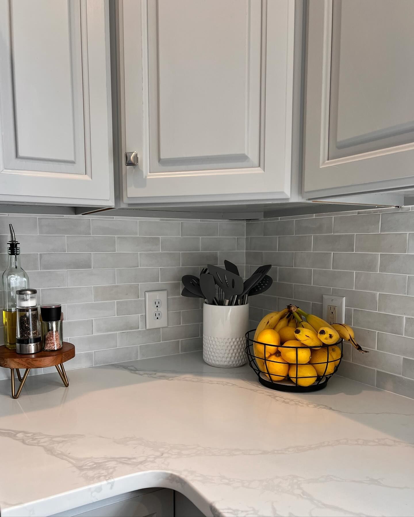Kitchen countertop with a wooden spice rack, a black wire basket of bananas, and a white utensil holder.