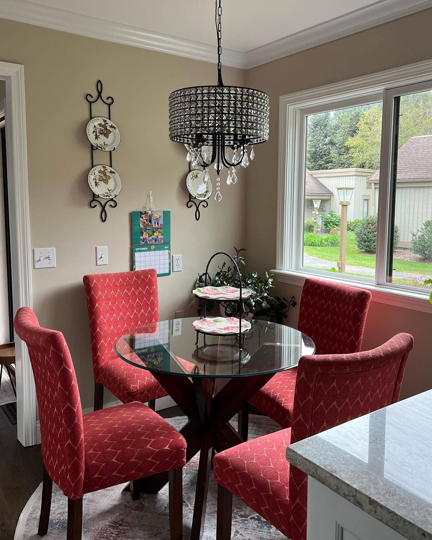 Dining area: round glass table, four red patterned chairs, crystal chandelier, decorative plates on the wall, and a window.