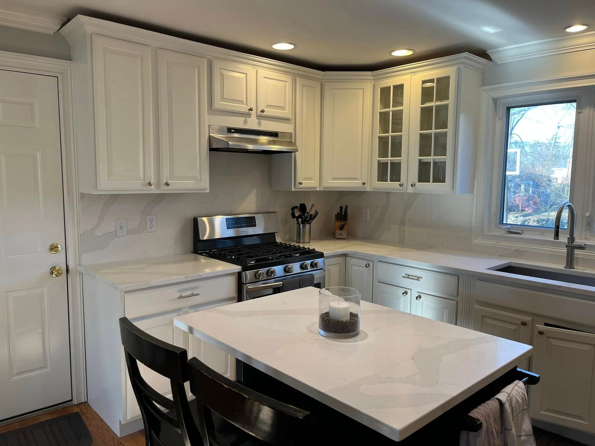 White kitchen with island, cabinets, stove, and sink. Window on right, black chairs at island.