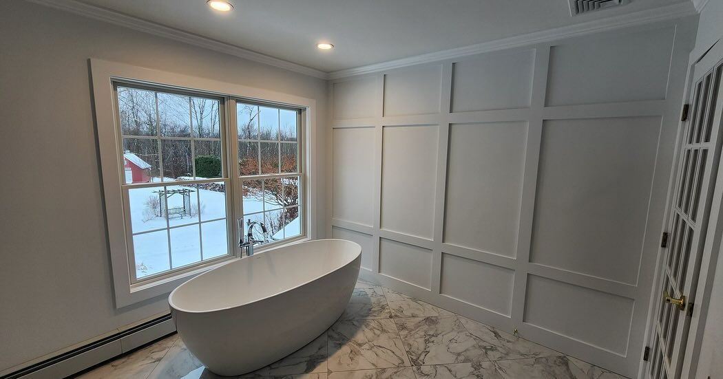 Bathroom with a white freestanding tub by a window overlooking a snowy landscape. White paneled walls.