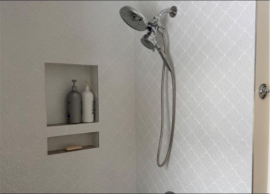 Shower with a built-in shelf containing shampoo bottles and soap, and a silver showerhead on patterned white tile.