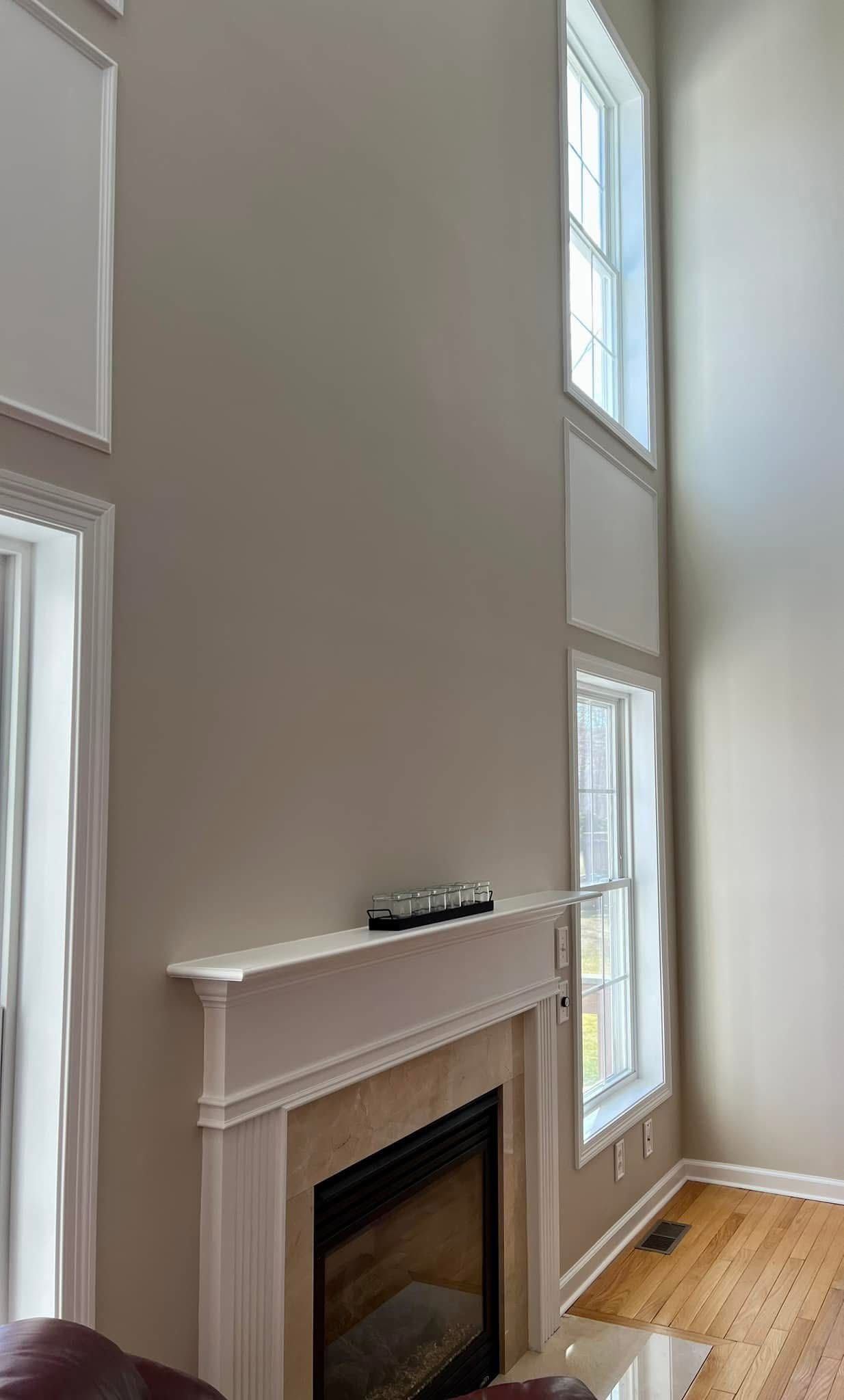 Fireplace with a white mantel and two-story tall windows. Light-colored walls and wood flooring.