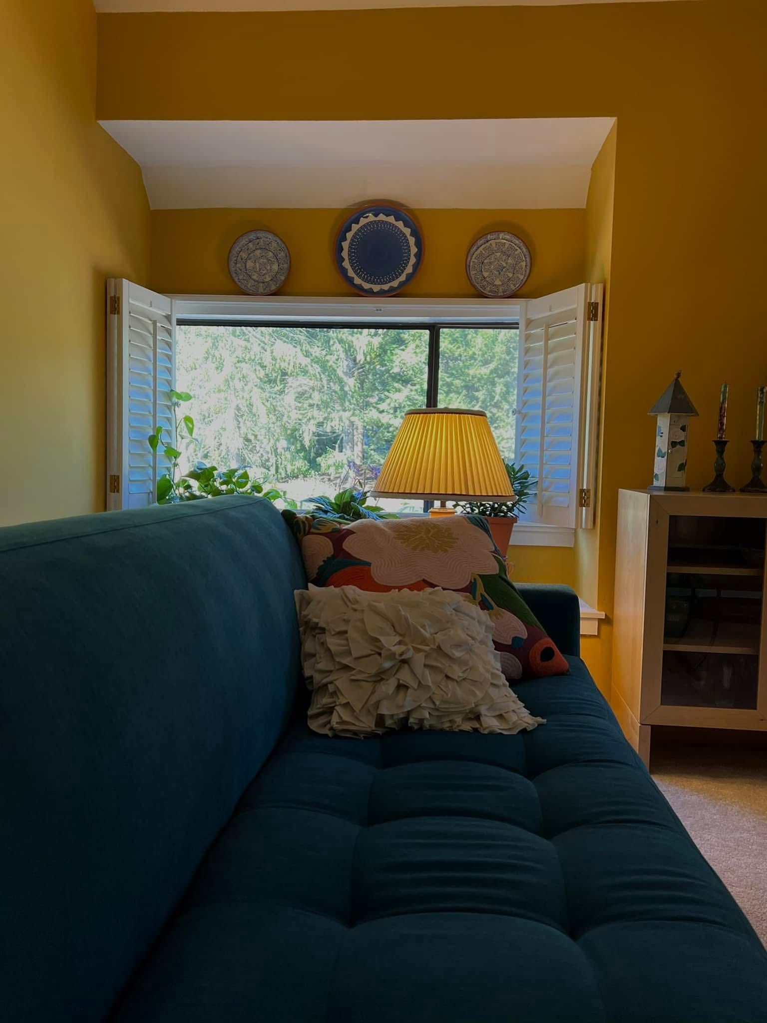 Teal sofa in front of a window with shutters, yellow walls, decorative plates, and a lamp.