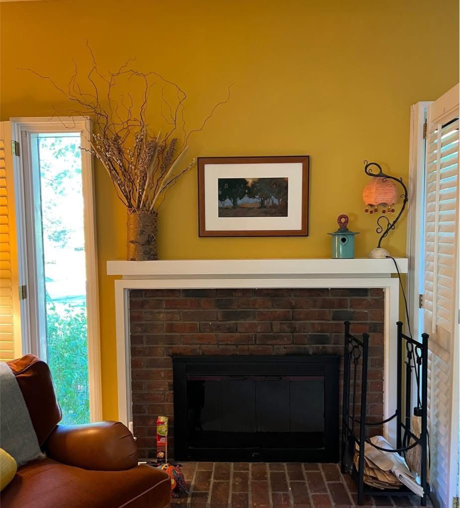 Fireplace with brick surround, white mantel, and yellow wall. Artwork, branches, and a lamp are on the mantel.