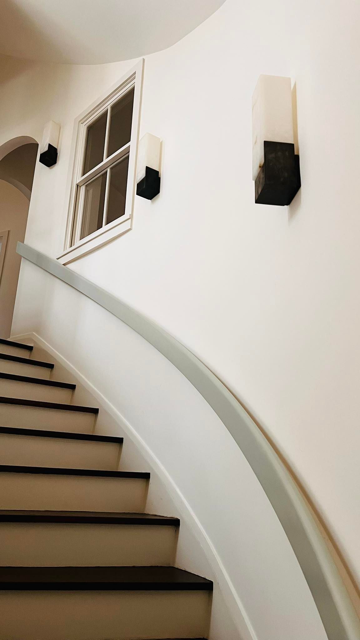 Staircase with white walls, curved railing, window, and wall sconces.