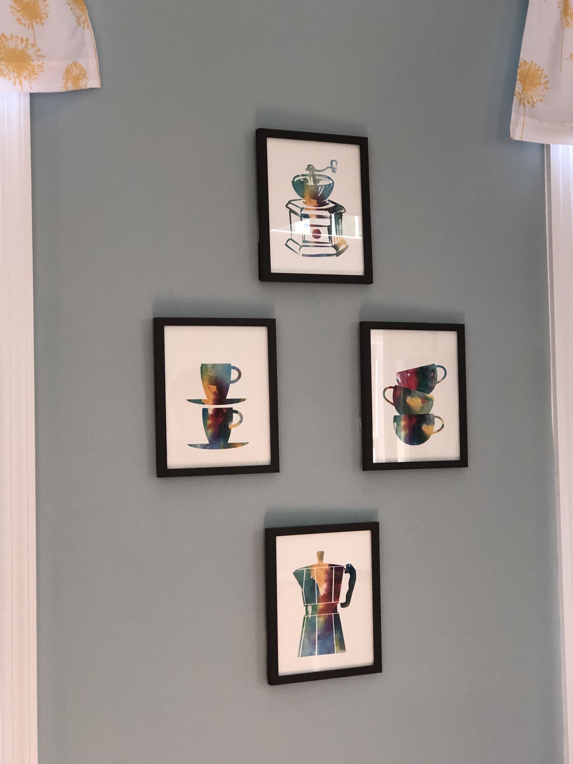 Four framed watercolor coffee art pieces on a light blue wall, with white window trim and curtains.