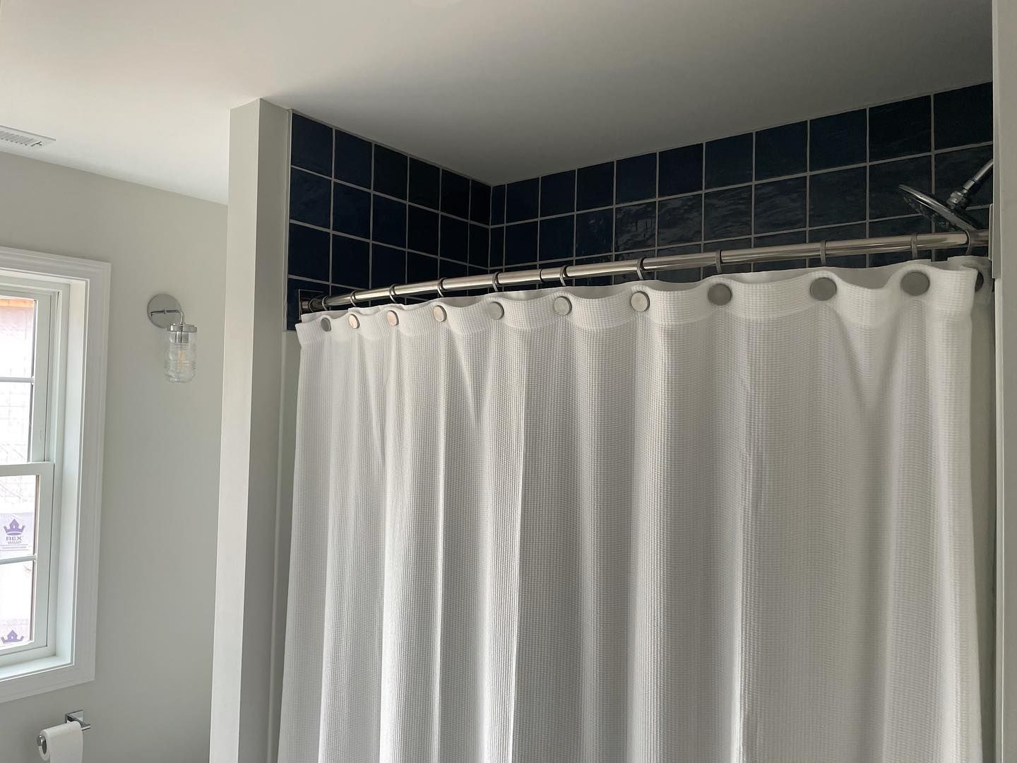 White shower curtain in a bathroom with dark blue tile and a window.