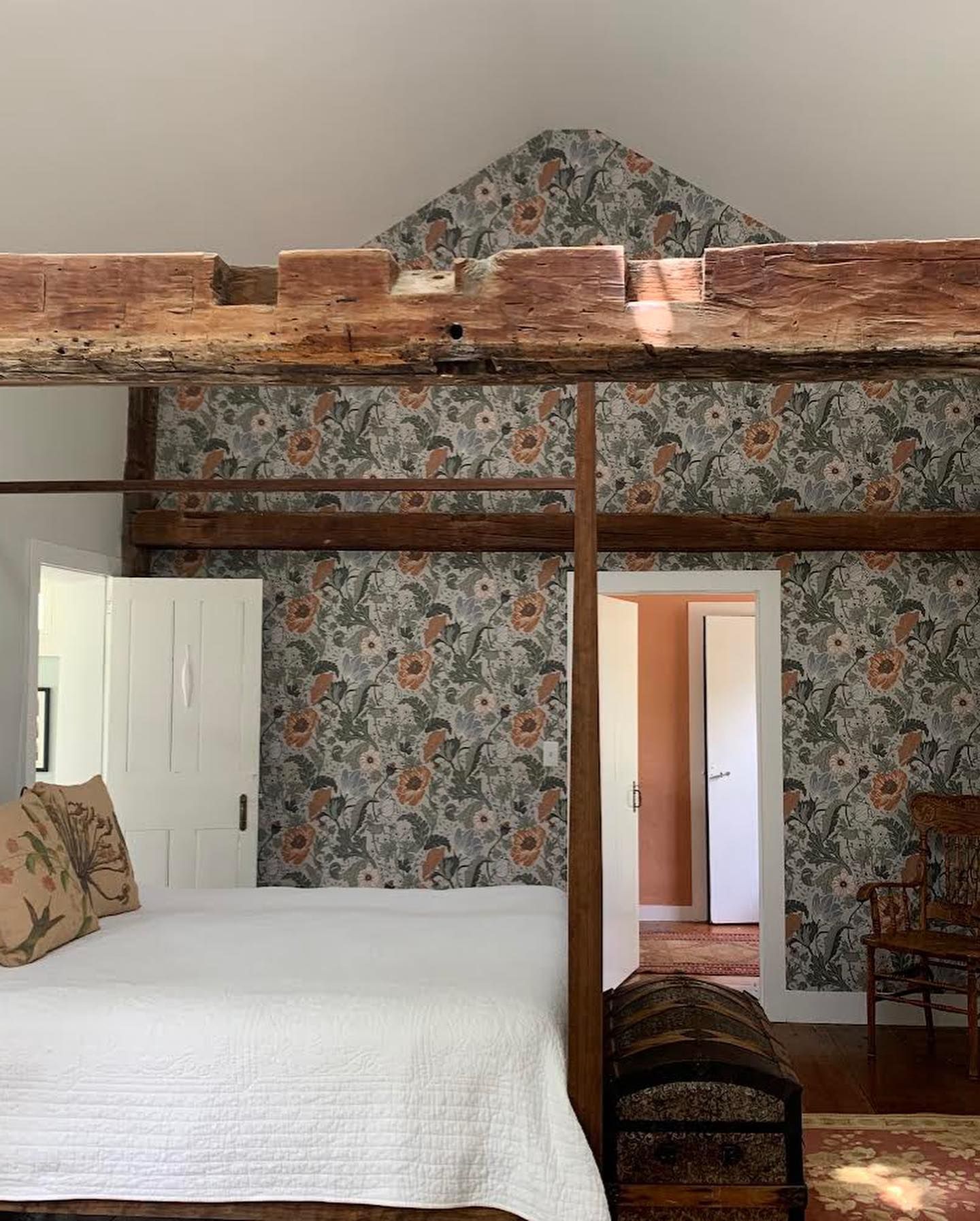 Bedroom with floral wallpaper and exposed wooden beams. A bed with white bedding is in the foreground.