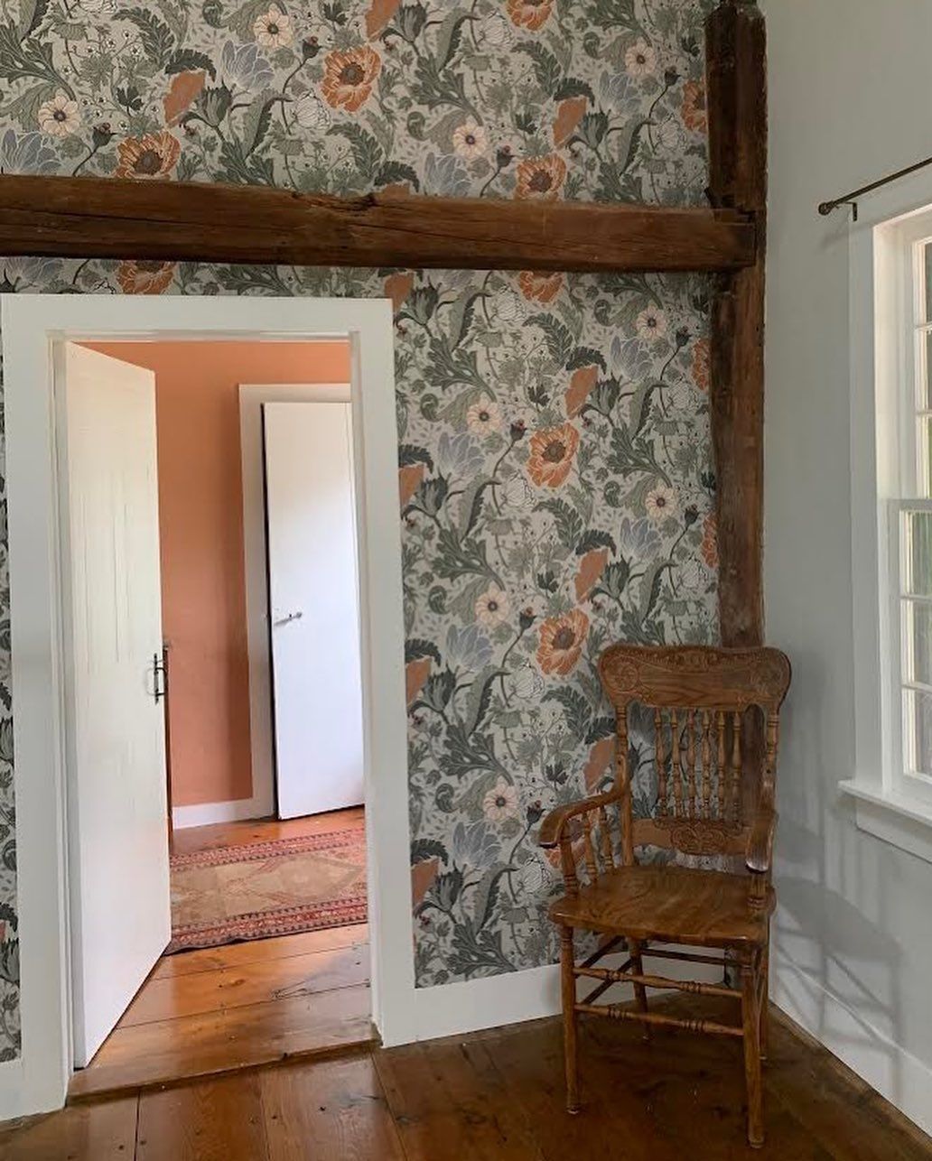 Room with floral wallpaper, wooden beams, chair, and doorway leading to another room.