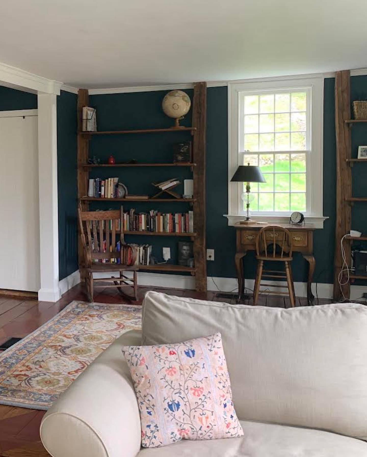 Living room with bookshelves, desk by window, and sofa with floral pillow; teal walls.