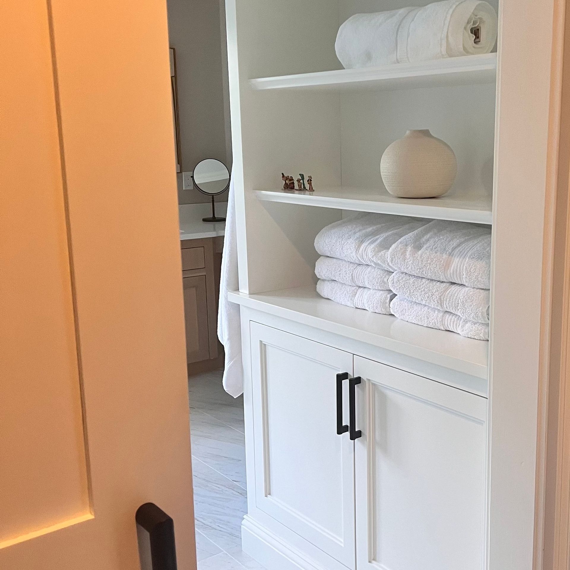 White built-in cabinet with towels, a vase, and a bathroom visible through an open door.