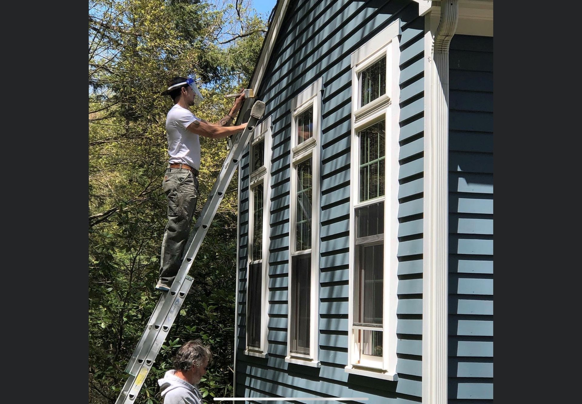 A man is standing on a ladder painting the side of a blue house.