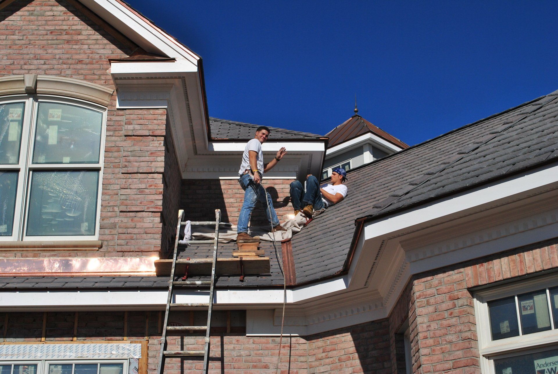 Two construction workers on a roof installing shingles on a brick building, using a ladder. Clear blue sky.