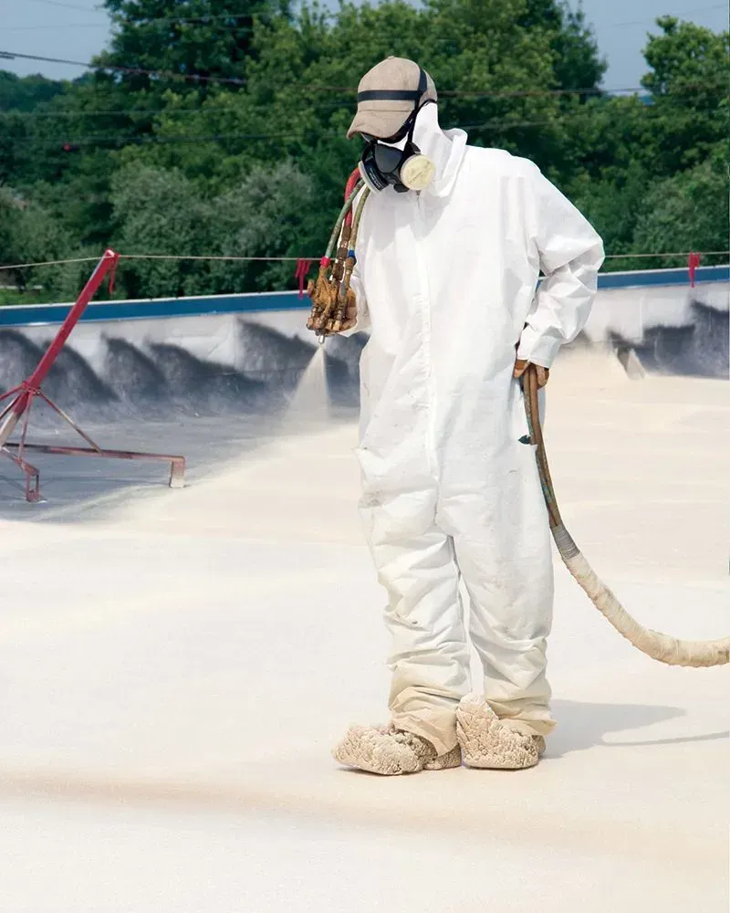 Worker in white protective suit spraying a rooftop surface with a hose and respirator mask.