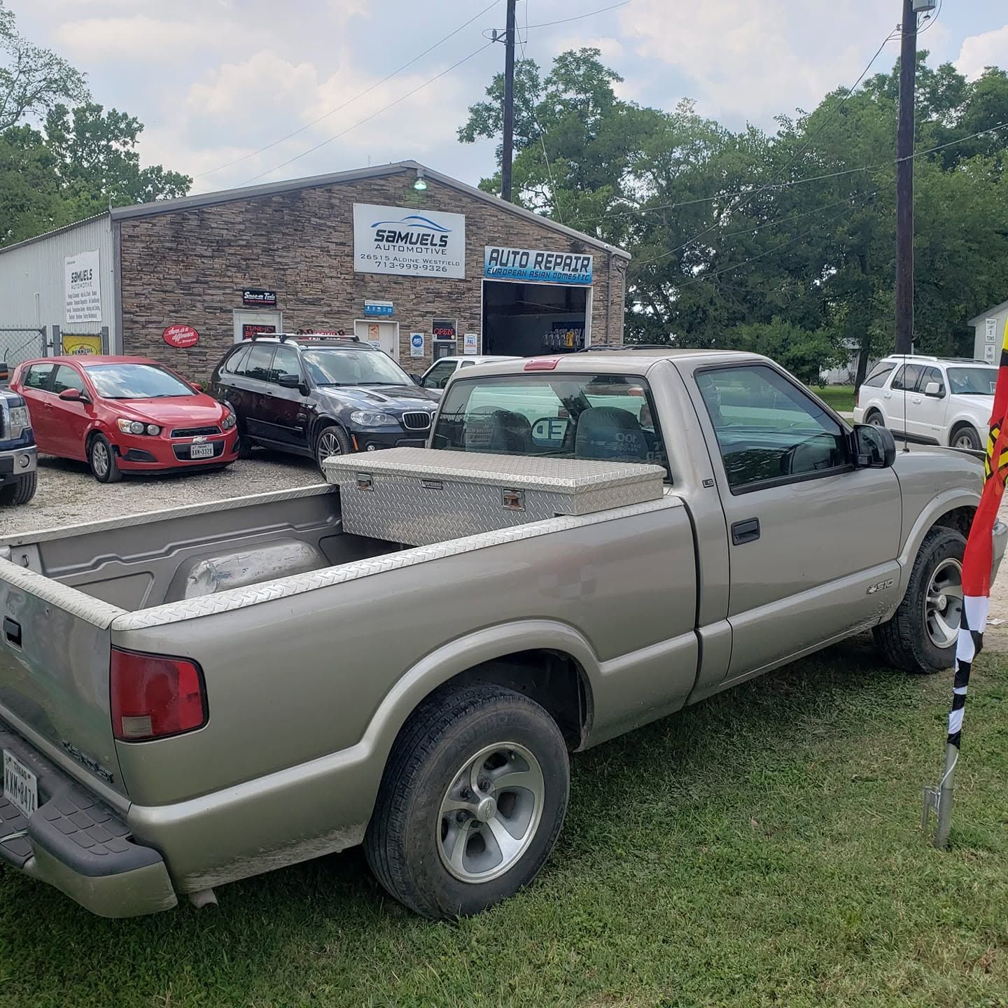 Silver pickup truck parked on grass in front of a brick auto shop with several cars nearby