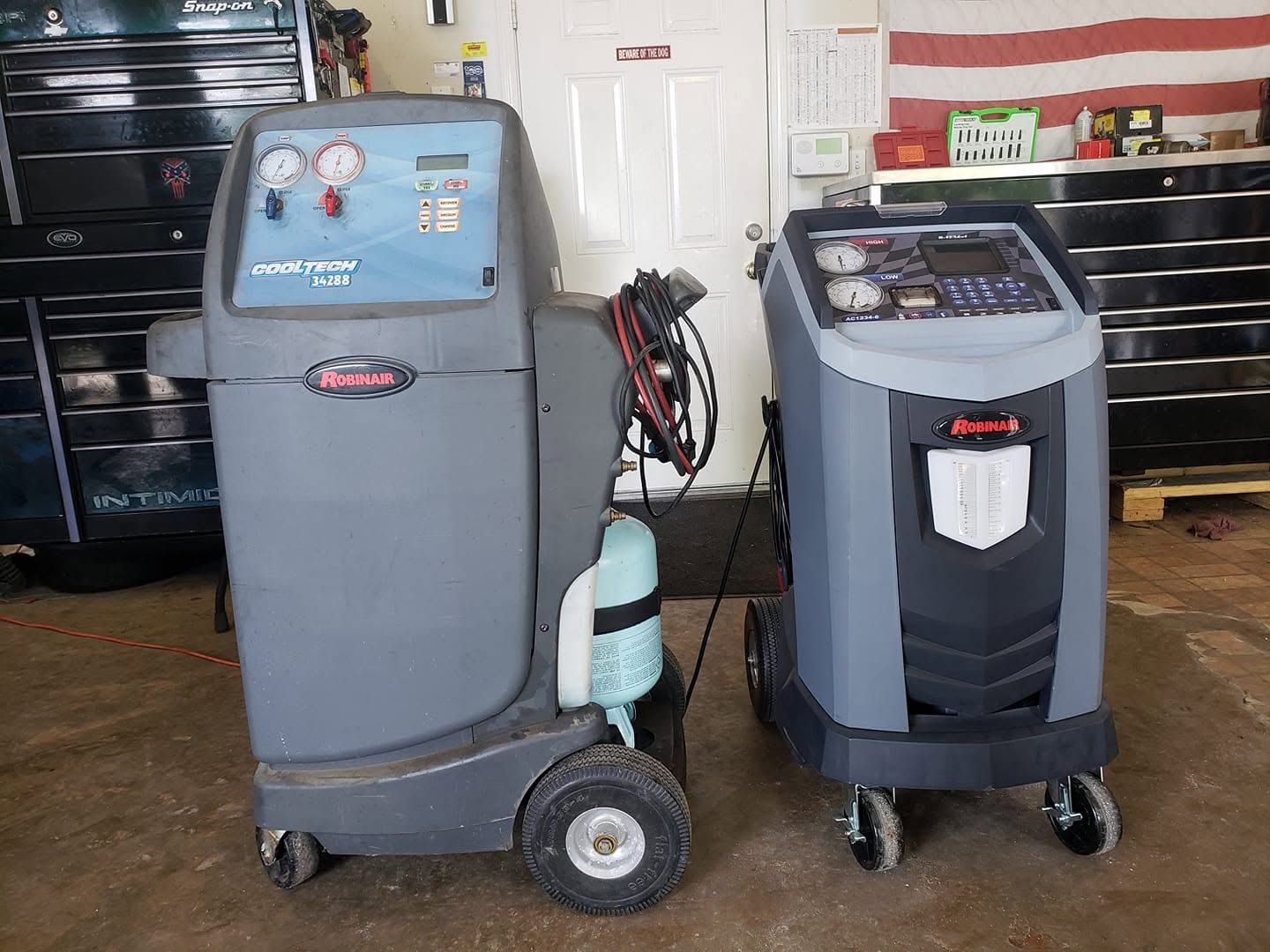 Two blue-gray floor scrubber machines in a garage, with a white door and tools in the background