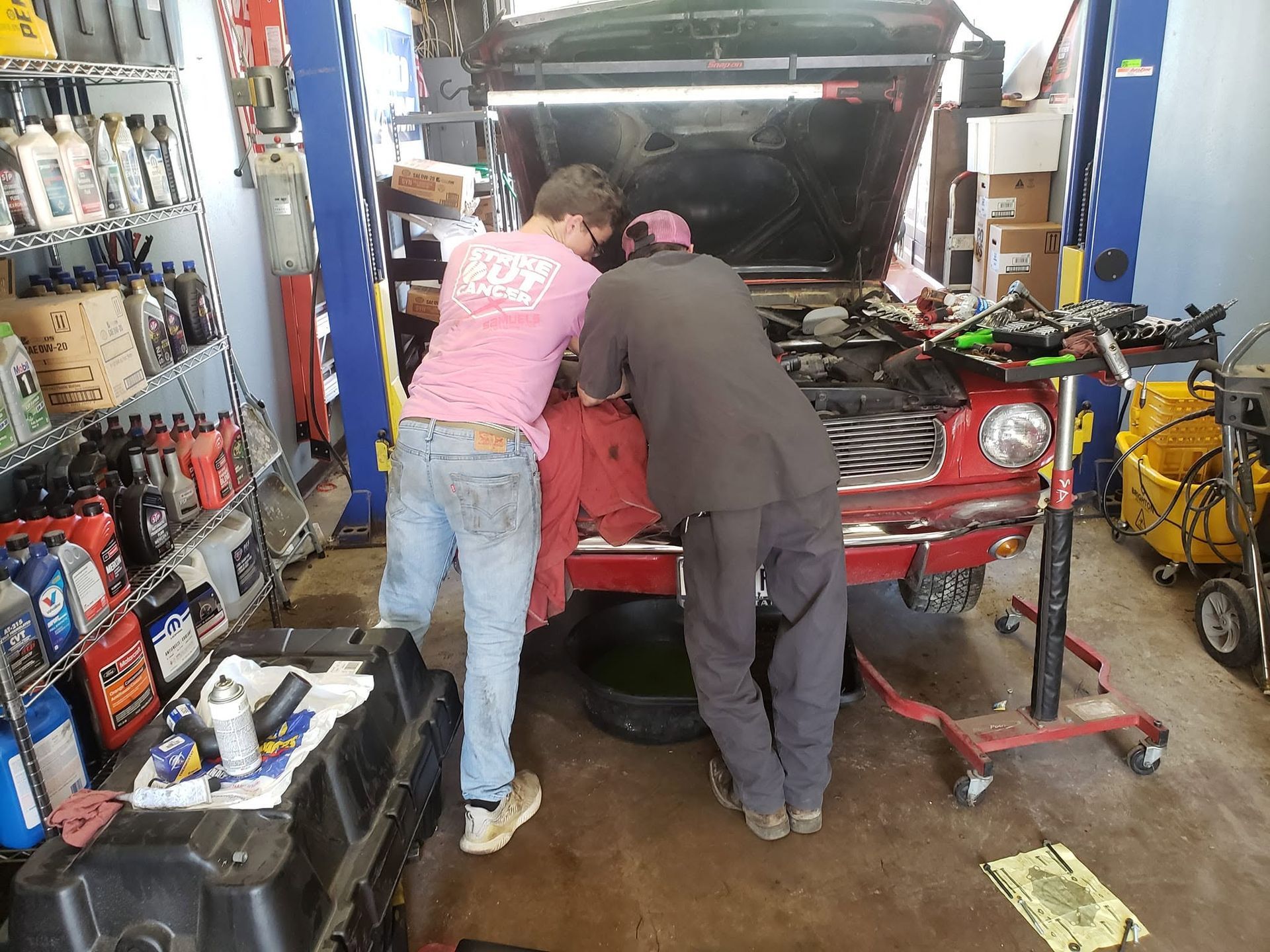 Two people working on a red car in a busy auto repair shop with tools and equipment around them