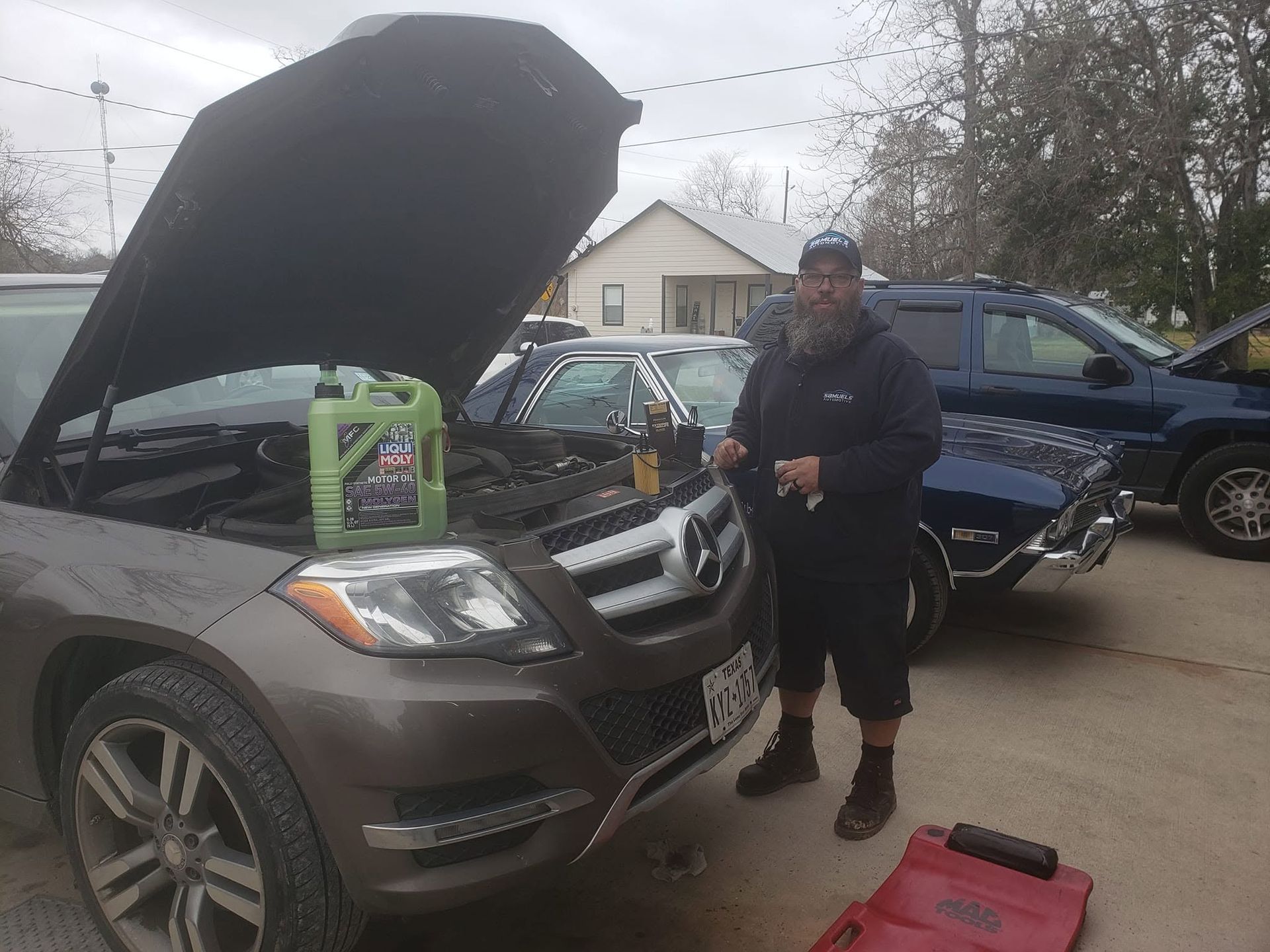 Man standing beside a gray SUV with its hood open, holding a tool in a driveway with other vehicles nearby