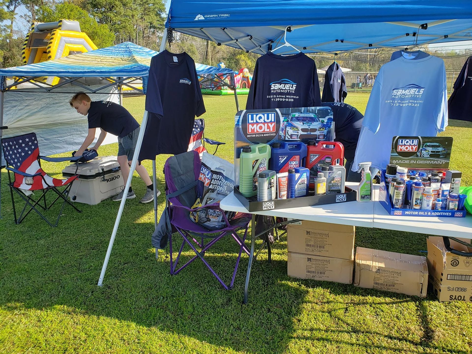 Outdoor vendor booth under blue canopy with navy shirts, camping chairs, and assorted merchandise on a grassy field