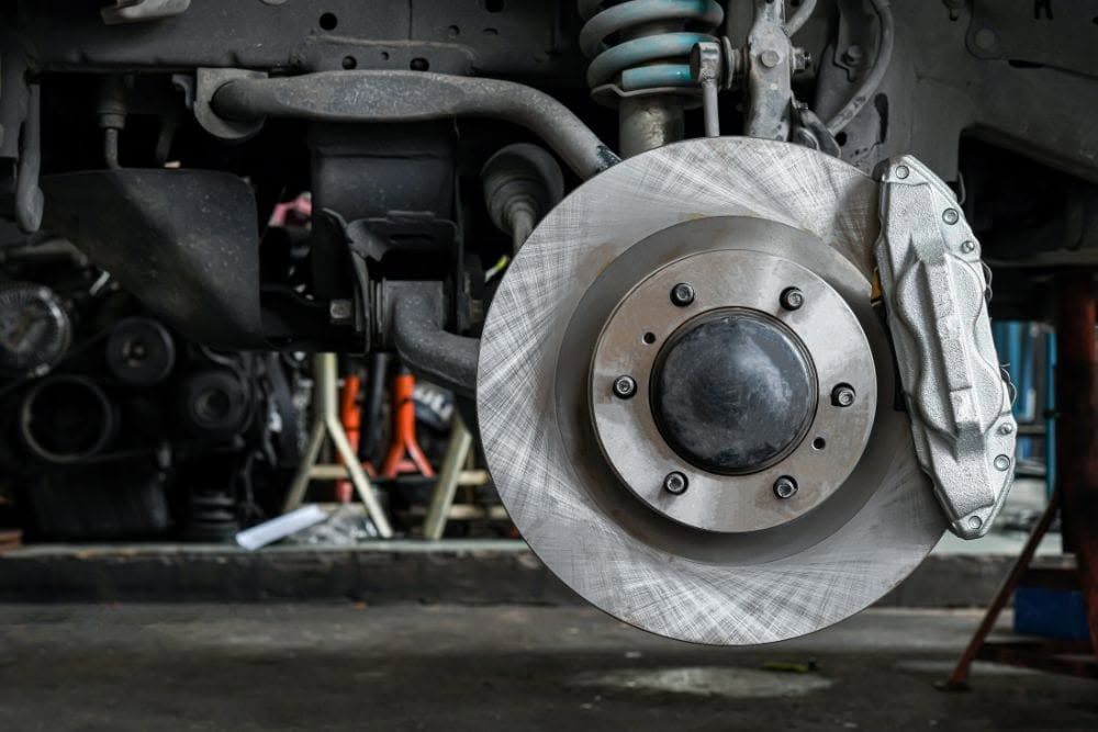 Close-up of a car’s front brake rotor and caliper in a garage, with the wheel removed.