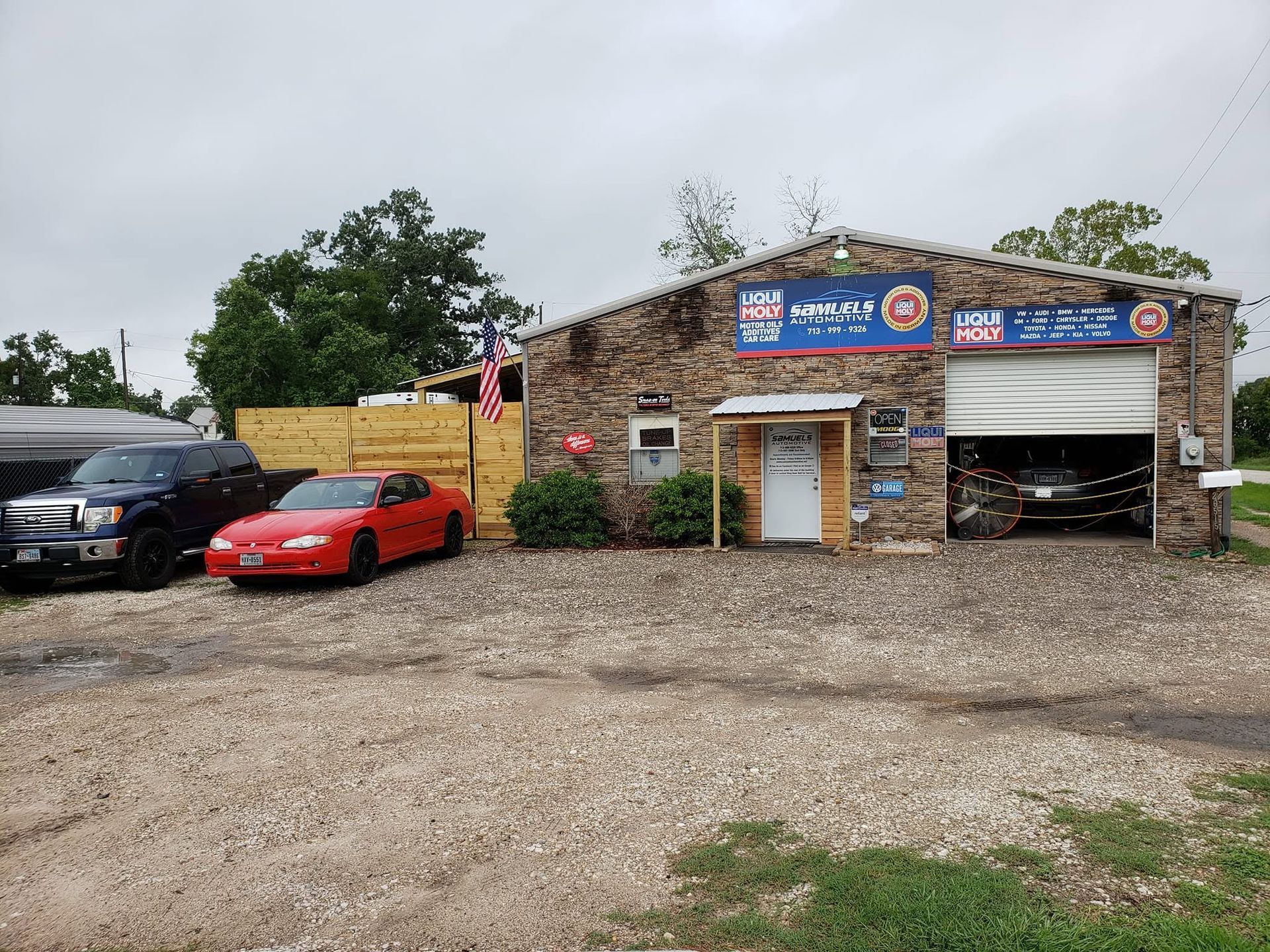 Auto repair garage with a red sports car and black SUV parked outside on a gravel lot.