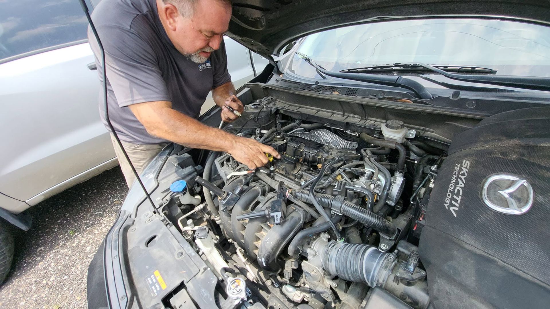 Mechanic working on a car engine in an outdoor parking lot beside a silver vehicle