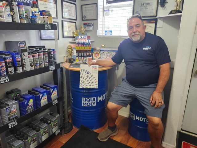 Man sitting in a small auto parts shop beside stacked motor oil containers and display shelves