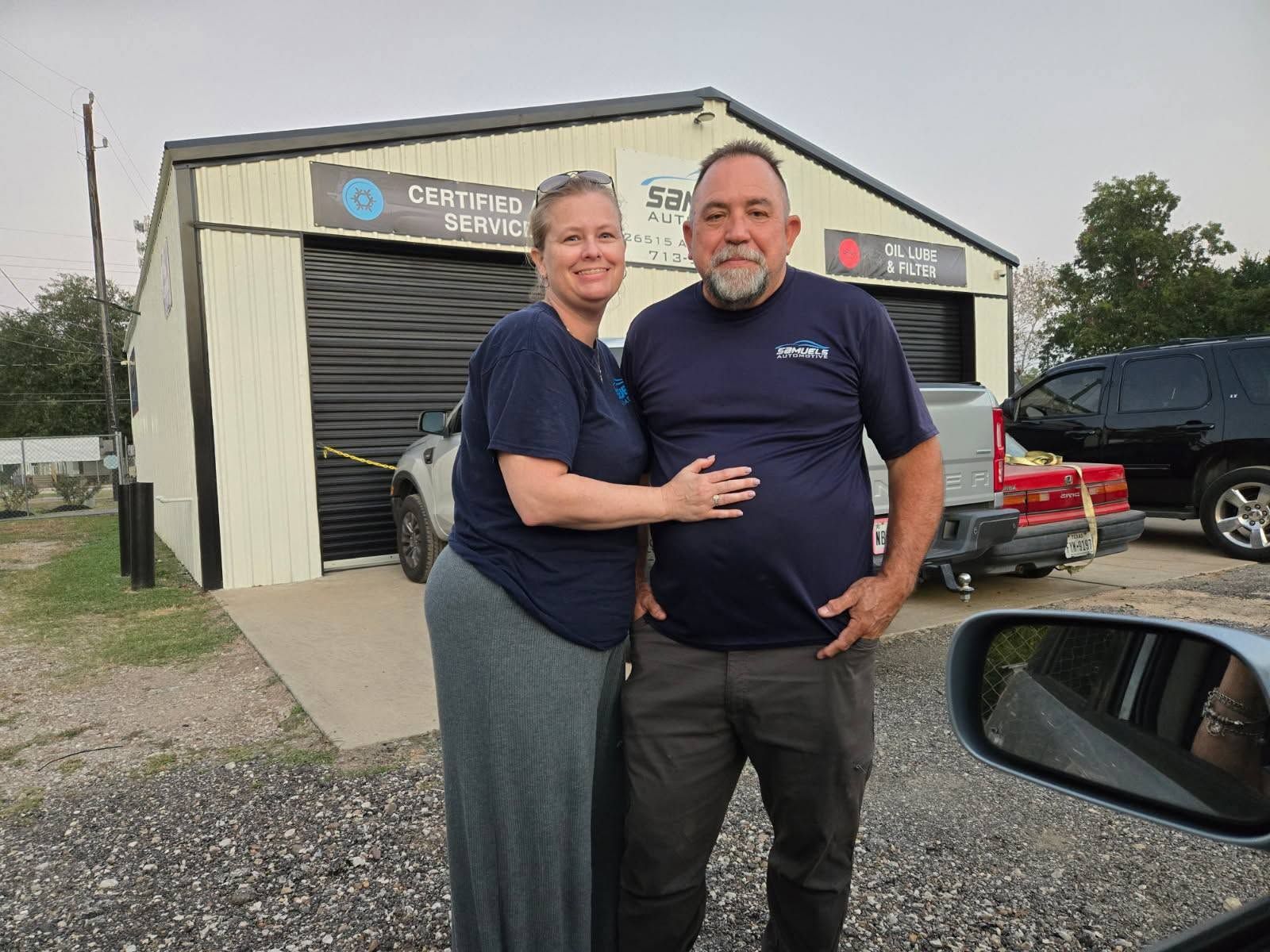 Two people standing and smiling in front of a beige building with garage doors and parked vehicles