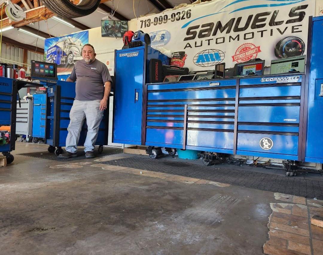 Mechanic standing beside blue tool chests in an auto shop with a “Samuels Automotive” sign behind him